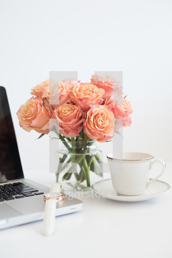 A laptop computer, white wristwatch, coffee cup and saucer and vase of peach roses on a white surface.