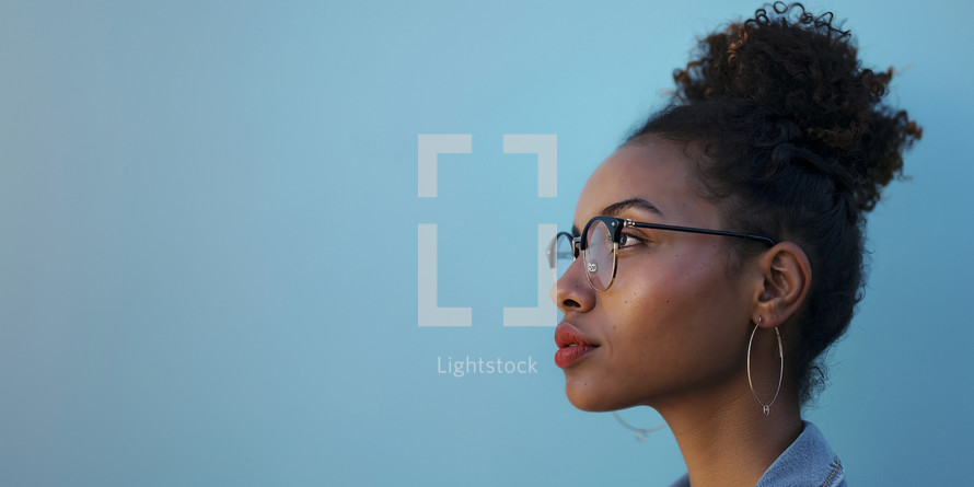 Young African American woman with glasses looking thoughtfully to the side, with a clear blue sky in the background.