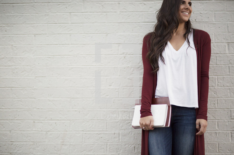 a young woman standing in front of a white wall holding a Bible and journal 