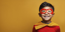 Delighted young boy in superhero costume with mask and cape, exuding excitement and joy on a golden yellow background.