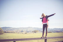 Woman balancing on a fence rail in a pasture with mountains.