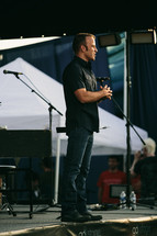 man at a microphone on stage at an outdoor worship service 
