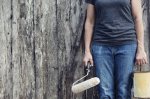 woman holding a paint bucket and paint roller in front of an old wood building.