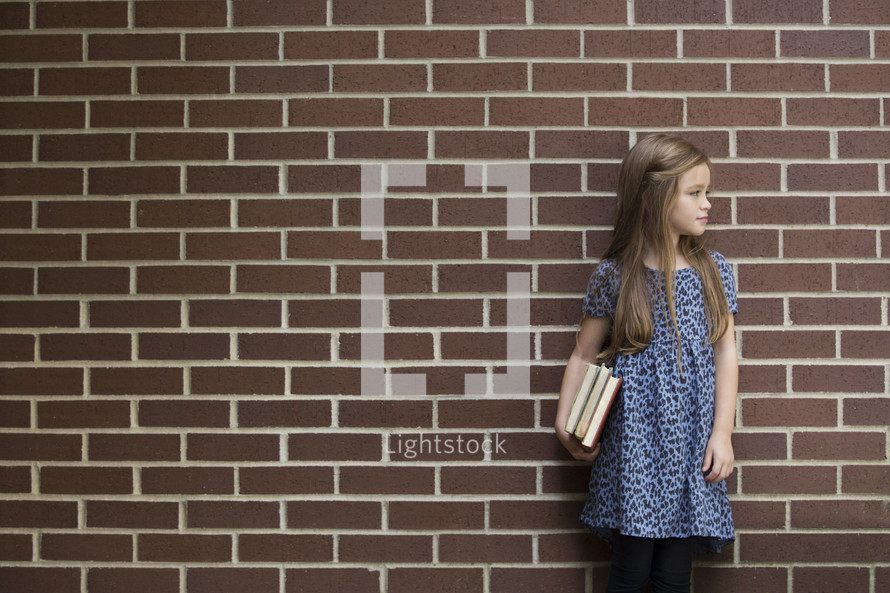 a young girl holding books in front of a brick wall 