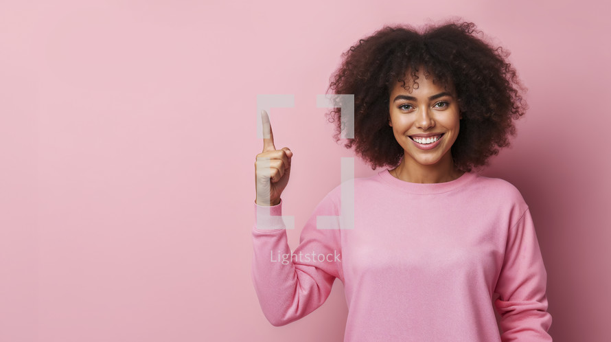 Enthusiastic young woman with curly hair, wearing a pink sweater and pointing upwards, with a bright smile on a pink background.