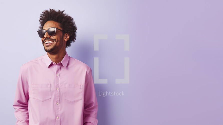 Smiling young man with curly hair wearing aviator sunglasses and a light pink shirt on a lavender background.