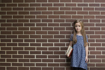 Little girl holding school books and standing against a brick wall.