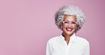 Radiant senior woman with curly gray hair and pink glasses smiling joyfully on a soft pink background.