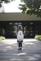 A little girl with a backpack looking at the front doors of a school.