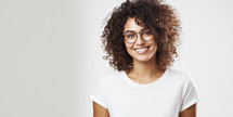 Confident young woman with curly hair and glasses, smiling in a white T-shirt, on a light background.