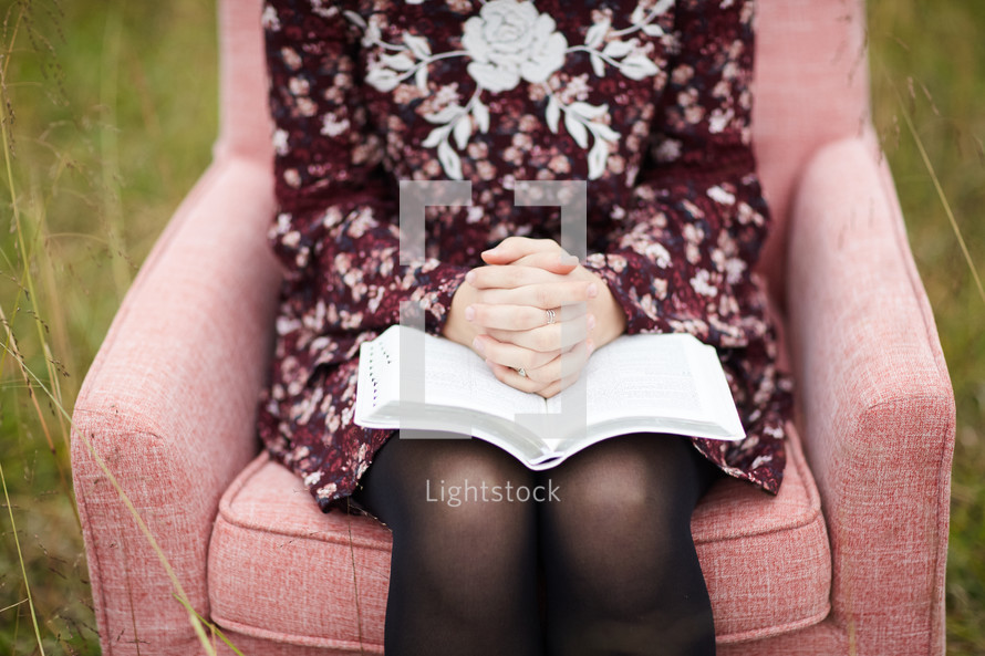 a young woman sitting in a chair in a field praying over a Bible 