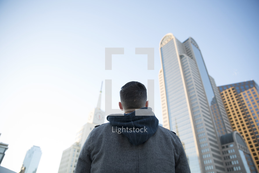 a man standing in a city looking up at the tall buildings.