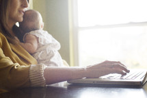 working mom, mother typing on a computer while holding her infant 