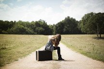 woman sitting on a suitcase on a dirt road with her face in her hands 