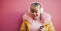Vibrant portrait of a cheerful young woman with Down syndrome, enjoying music on white headphones, against a pink background, wearing a yellow jacket with a pink fur collar.