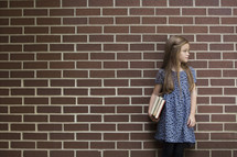 a young girl holding books in front of a brick wall 