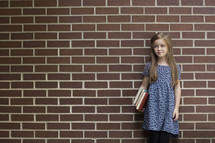 a young girl holding books standing in front of a brick wall 