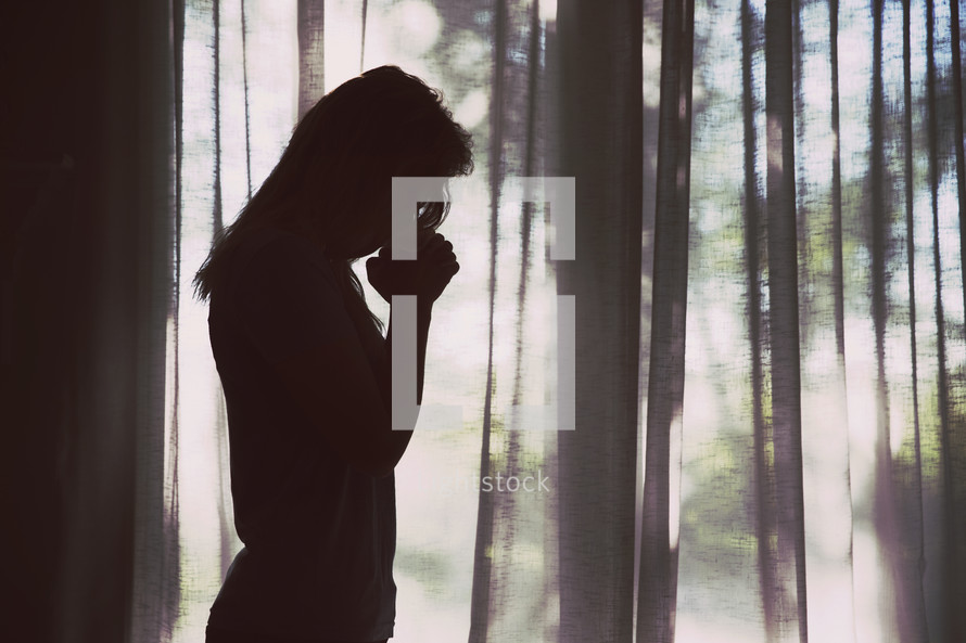 silhouette of a woman with head bowed in prayer standing alone in a forest 
