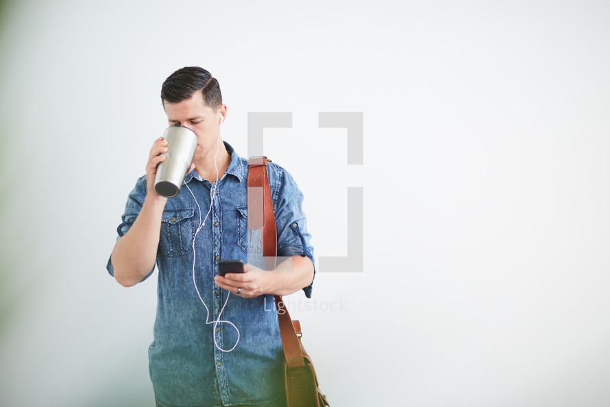 a man standing holding a messenger bag listening to earbuds and drinking coffee 