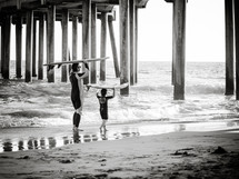 father and son carrying surfboards under a pier on a beach 