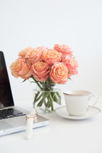 A laptop computer, white wristwatch, coffee cup and saucer and vase of peach roses on a white surface.