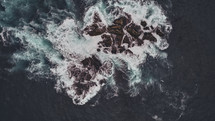 aerial view over waves crashing into a rocky island 