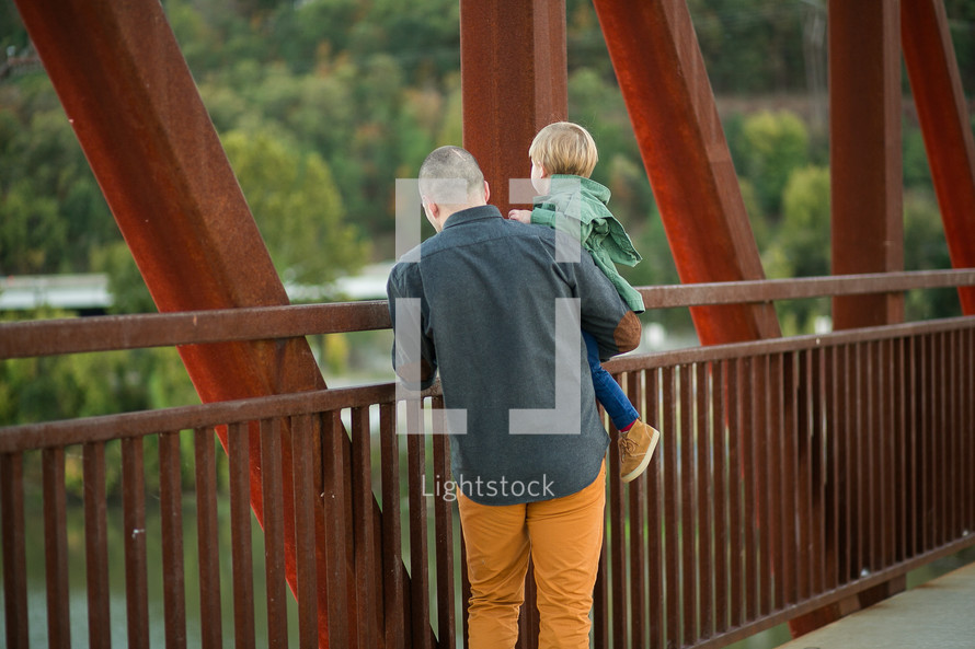 a father holding his son as they look over a bridge railing 
