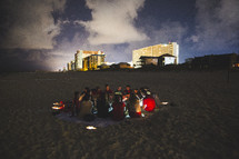 group of people sitting on a blanket in the sand on a beach at night 
