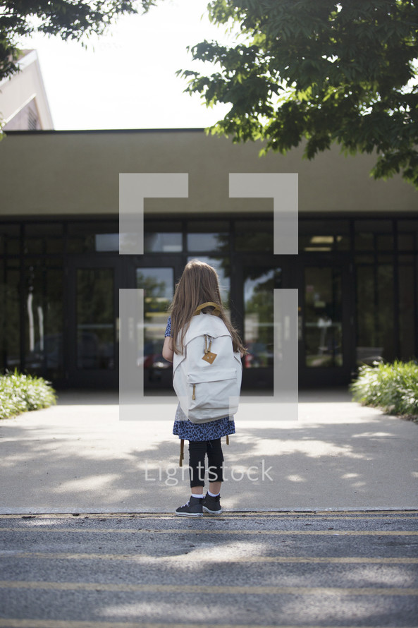 A little girl with a backpack looking at the front doors of a school.
