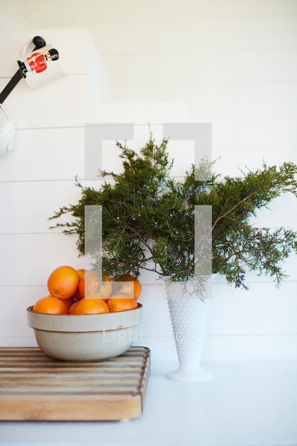 bowl of oranges and vase of evergreen branches 