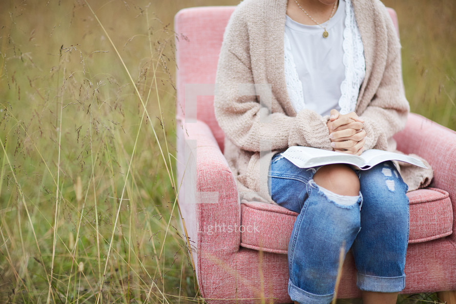 a woman sitting in a chair in a field with praying hands over the pages of a Bible 
