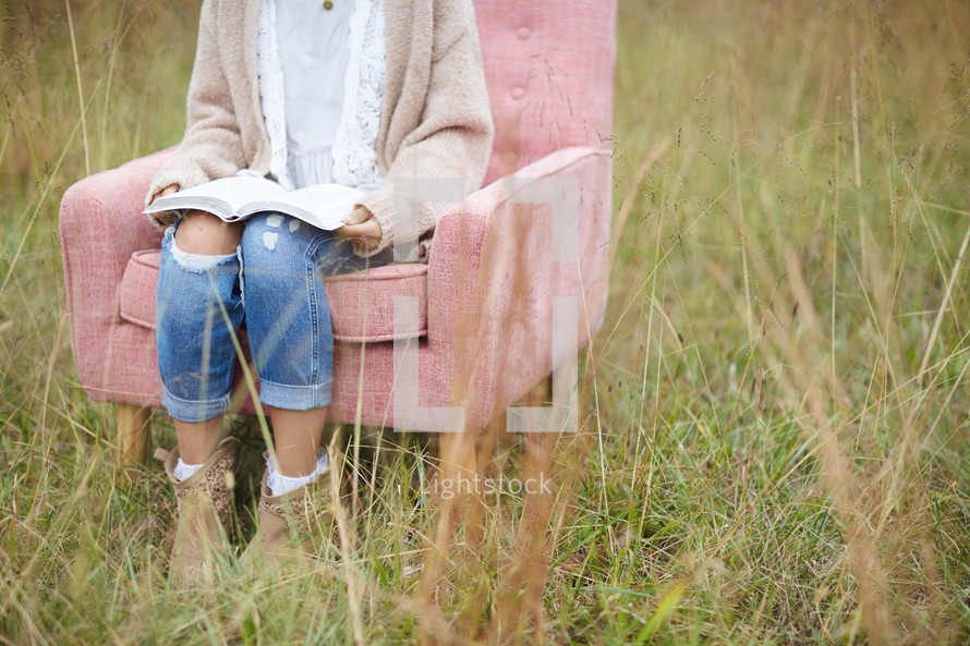 a woman sitting in a chair reading a Bible in a field 