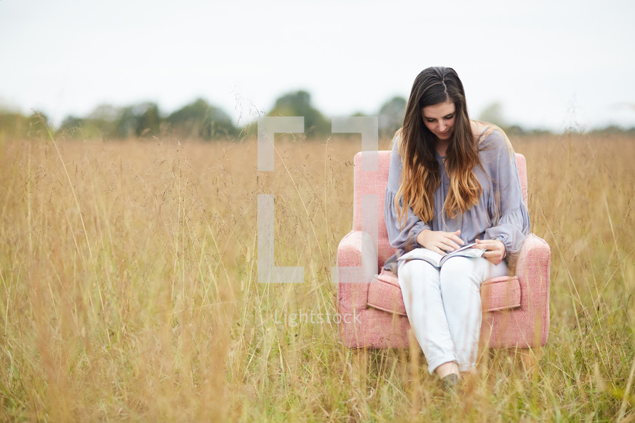 a young woman sitting in a chair in a field reading a Bible 