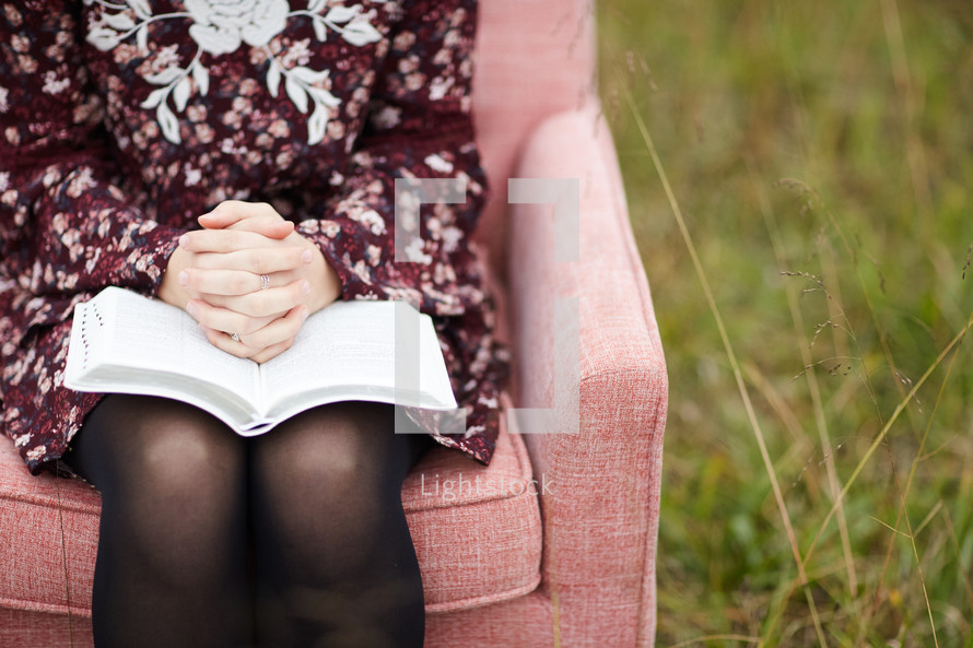 a young woman sitting in a chair in a field with praying hands over the pages of a Bible 