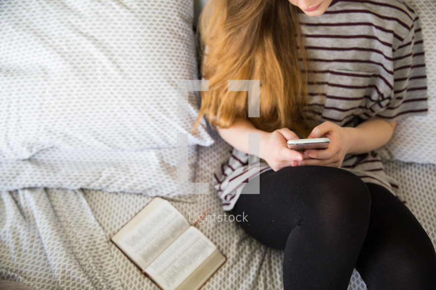 girl texting sitting on a bed and an open Bible 