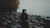 a young woman in a trench coat and hat standing on a rocky shore 