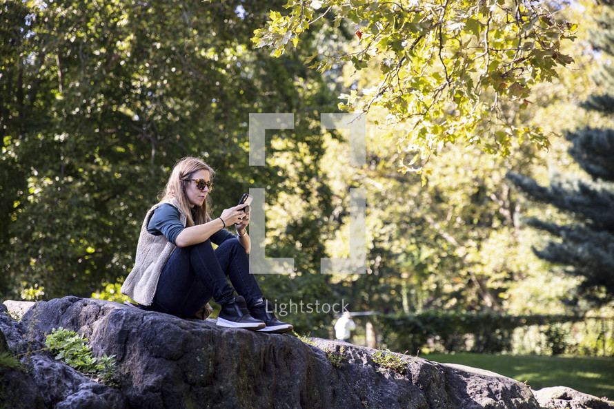 Woman sitting on a rock in a park while looking at her phone