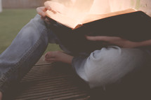 A barefooted person in jeans sitting outdoors with a Bible in his lap.