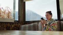 Pretty woman sitting on the cafe terrace on the background of winter mountain landscape, using a smartphone. Woman tourist uses internet on smartphone on restaurant in mountain.Amazing mountains view.
