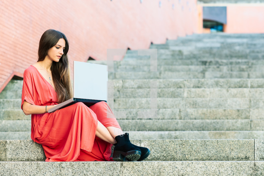 young woman sitting on outdoors stairs wearing red dress with a computer 