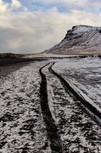 Snow covered black sand beach on the Snaefellsnes peninsula in Iceland with tire tracks leading into the distance toward a cliff.