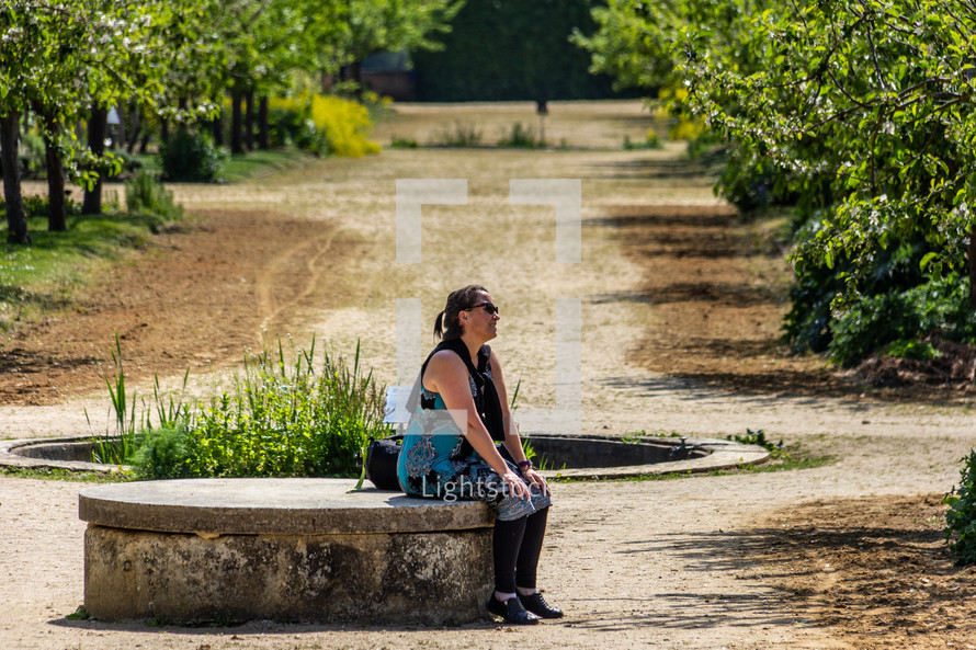 A woman sits contemplatively on a concrete bench in a grove of trees in garden near a fountain