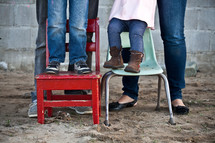 The legs of a man and woman standing behind a boy and girl who are standing in chairs placed in the dirt