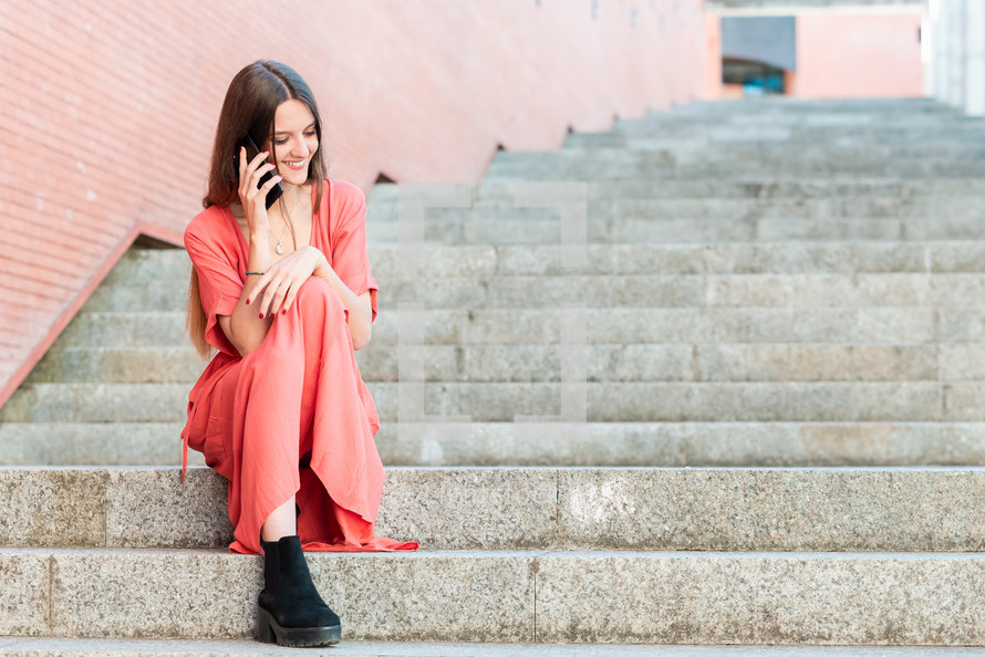 young woman sitting on outdoors stairs wearing red dress talking on a cellphone 