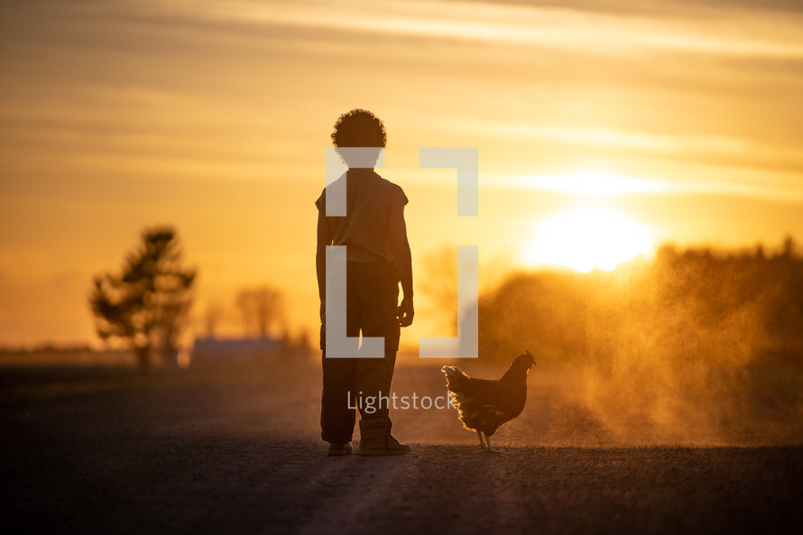 a boy standing on a dirt road at sunset next to a chicken 