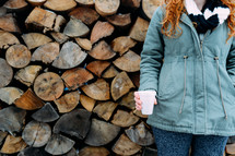 woman in a winter coat standing next to a stack of firewood 