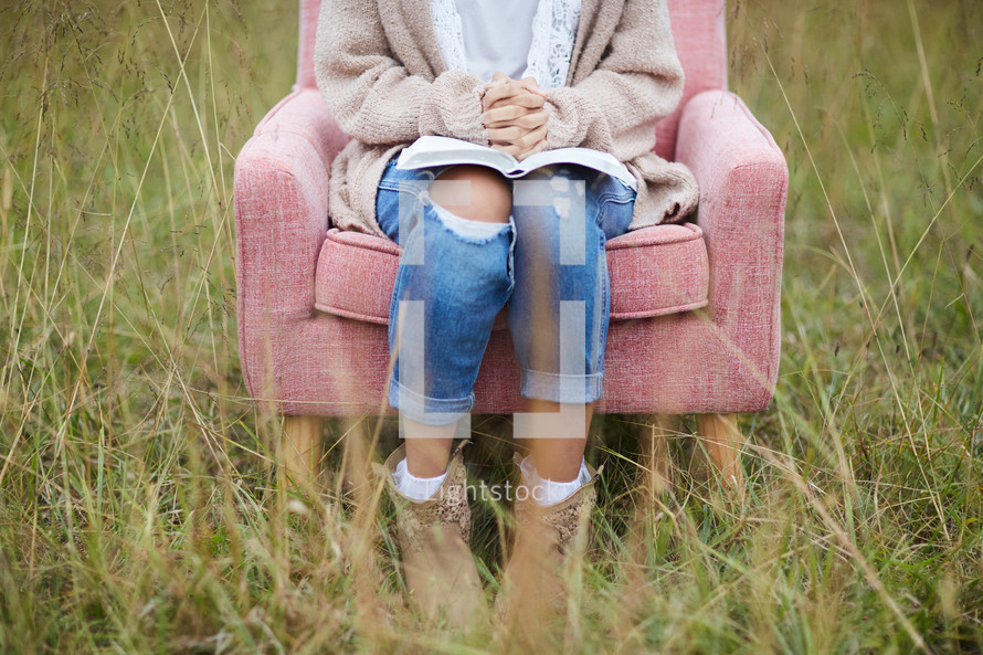 a woman sitting in a field with praying hands over the pages of a Bible 
