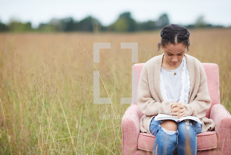 a woman sitting in a chair with praying hands over a Bible in a field 