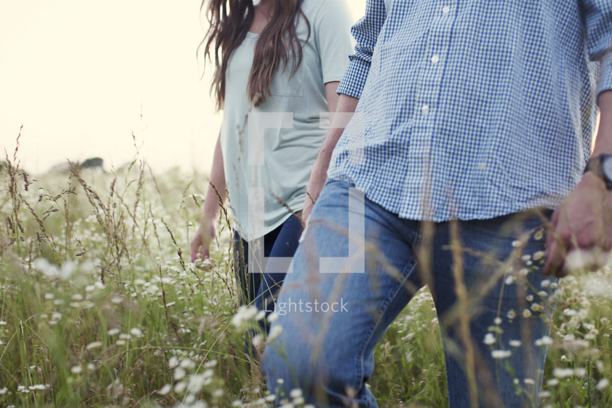 a couple walking holding hands through a field of tall grass 