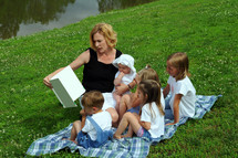 Woman reading a book to a group of children, sitting on a blanket on a hillside